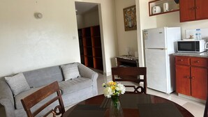 A compact living space with a grey sofa, wooden chairs, a small dining table with a vase of flowers, a kitchen area with a microwave and refrigerator, and a wooden cabinet above the sink. - St. Lawrence Suites - Suite Three (Oistins)