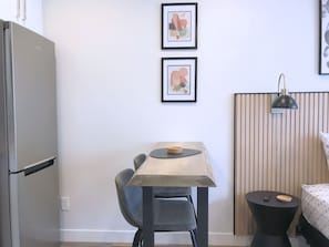 Una habitación compacta con kitchenette, una pequeña mesa de comedor, una silla, una cama con cubrecama estampado y dos cuadros en la pared.