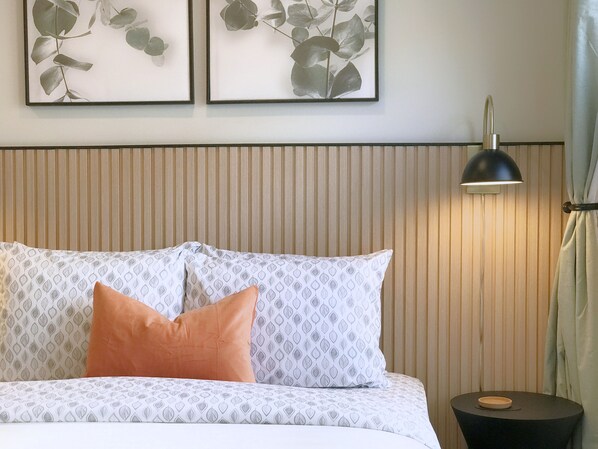 Una cama bien tendida con ropa de cama estampada, una única almohada naranja y una mesita de noche con una lámpara.