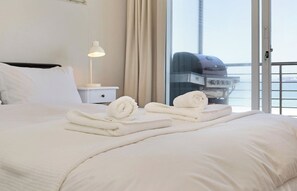 A hotel room with a bed, a nightstand with a lamp, and a view of the sea through a large window.