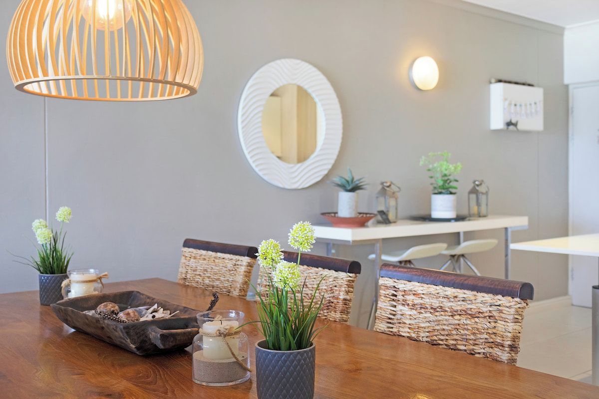 Uma sala de jantar com mesa de madeira, cadeiras de vime, espelho decorativo e luminária pendente.