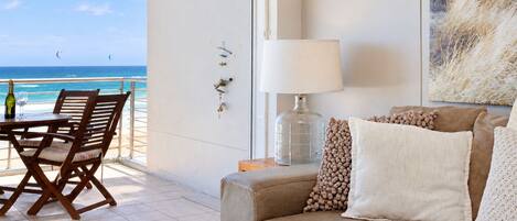 A balcony with a view of the ocean, a table with chairs, a sofa with cushions, a lamp, and a wall-mounted light fixture.