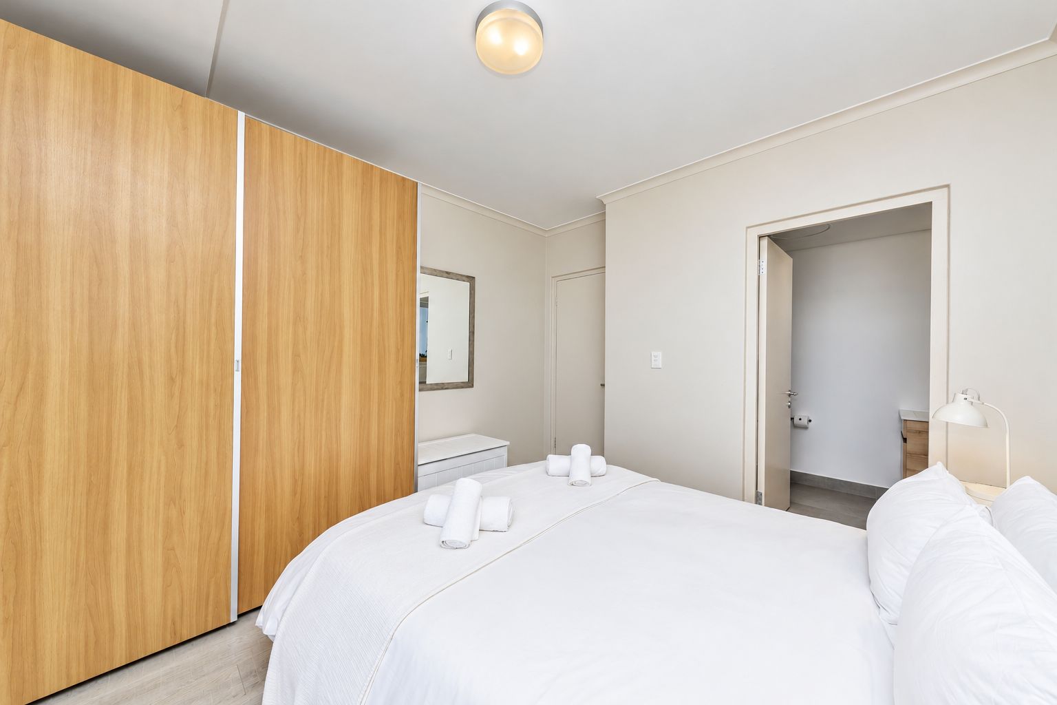 A hotel room with a bed, a wooden wardrobe, a mirror, a bathroom, and a nightstand with a lamp.
