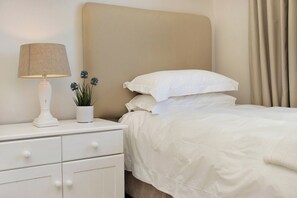 A neatly made bed with white bedding, a bedside lamp, and a small potted plant on a white nightstand. - Beachfront Apt - Kite Beach + table mtn view (Cape Town)