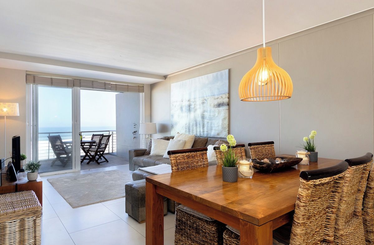 A modern living room with a wooden dining table, wicker chairs, and a large window overlooking the sea.