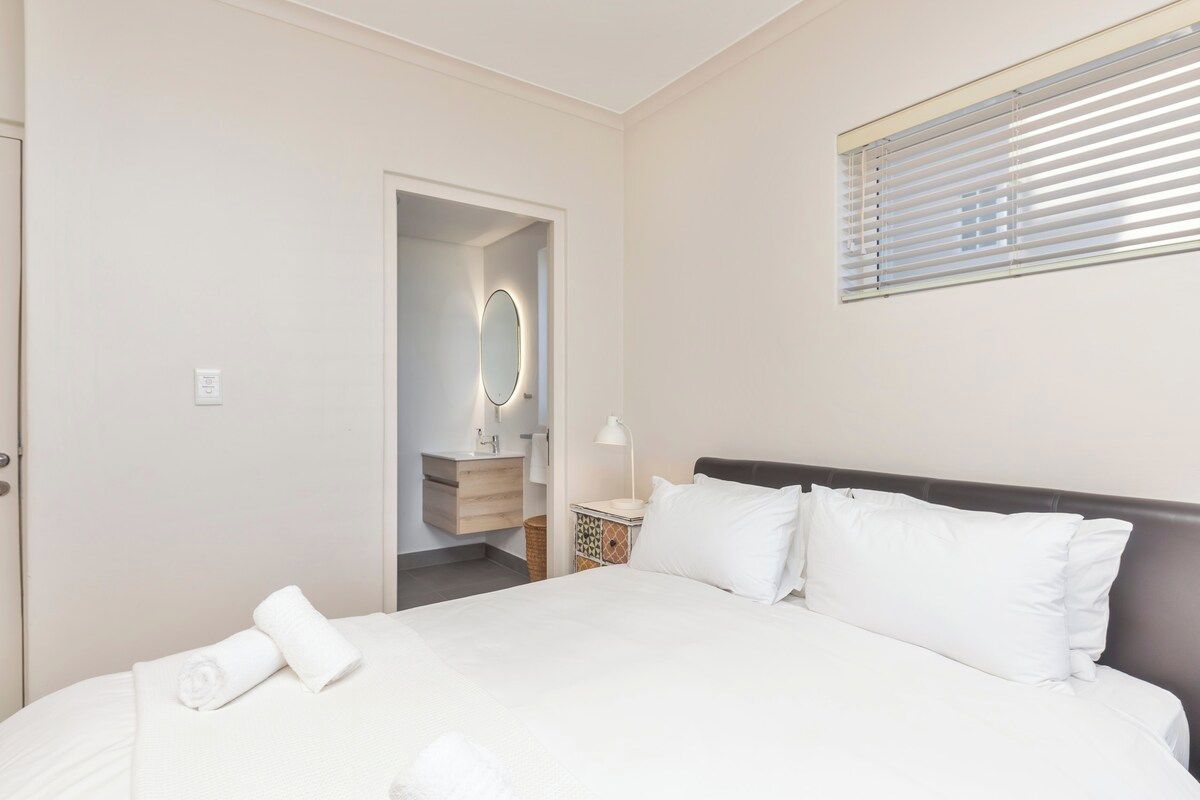 Habitación de hotel con cama, mesita de noche, espejo y ventana con persianas.