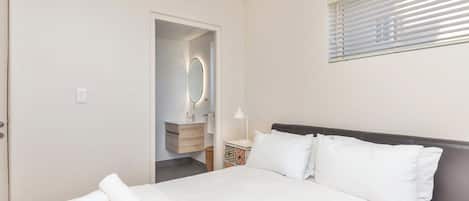 Habitación de hotel con cama, mesita de noche, espejo y ventana con persianas.