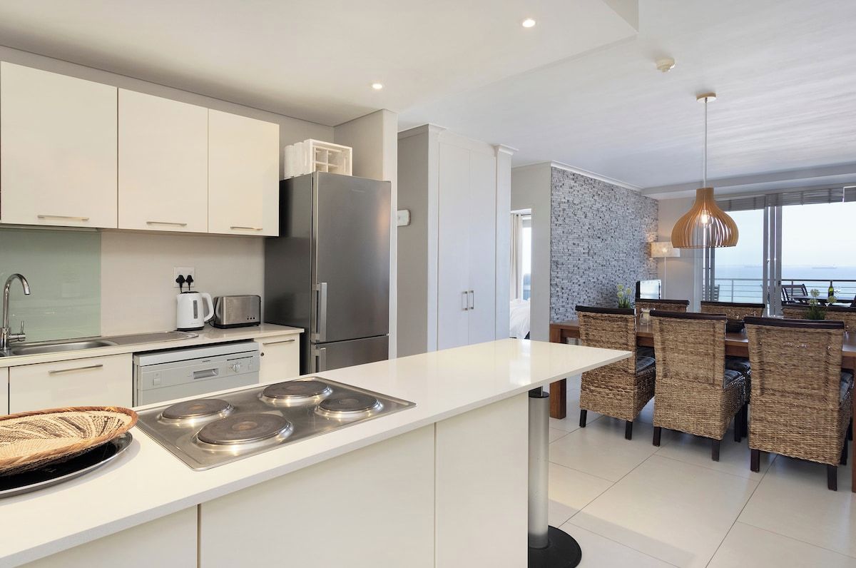 A modern kitchen with white cabinetry, stainless steel appliances, and a dining area with wicker chairs.