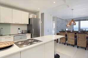 A modern kitchen with white cabinetry, stainless steel appliances, and a dining area with wicker chairs. - Beachfront Apt - Kite Beach + table mtn view (Cape Town)