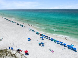 A beach with rows of blue and red umbrellas, white sand, and clear turquoise water. - Spring sale! Luxury listing! Walk to the beach! (Destin)