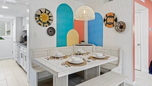 Un comedor con una mesa blanca dispuesta para cuatro personas, rodeado de coloridas obras de arte en la pared y una lámpara colgante.