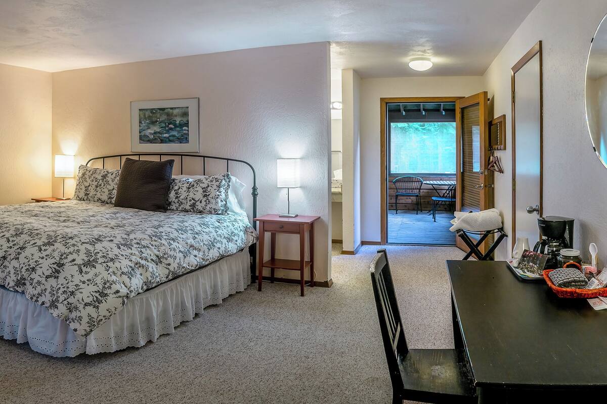 A bedroom with a bed, bedside lamps, a small table, and a chair. There is a door leading to a balcony with outdoor seating.