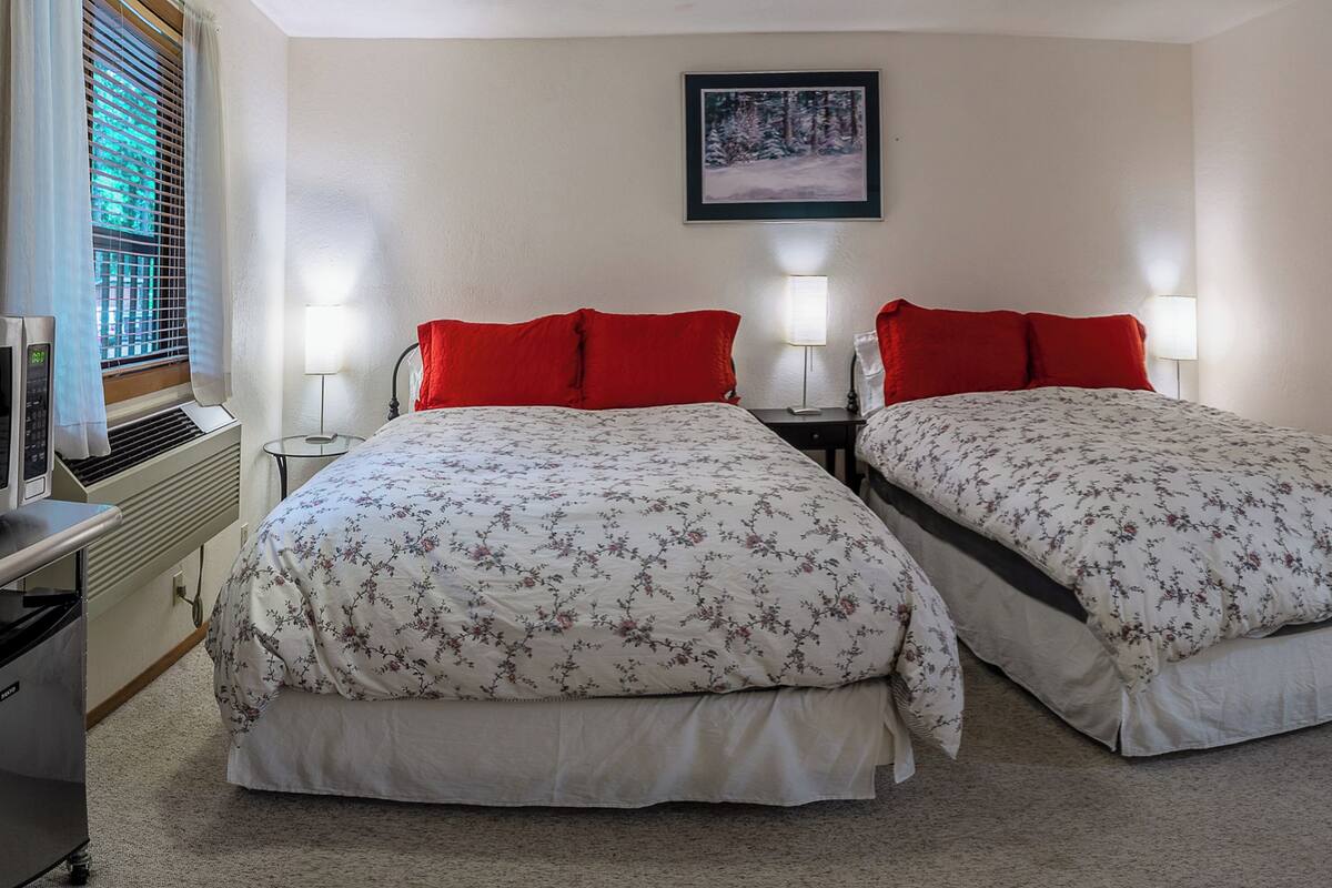 A double room with two beds, each with a floral bedspread and red pillows. There is a small bedside table with a lamp, a heater, and a window with blinds.