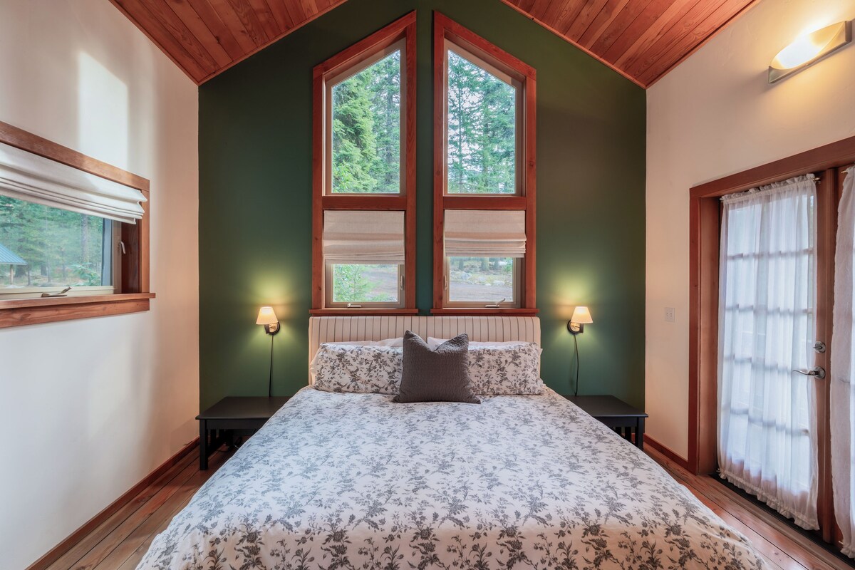 A bedroom with a large bed, two bedside lamps, a window with blinds, and a door with curtains.