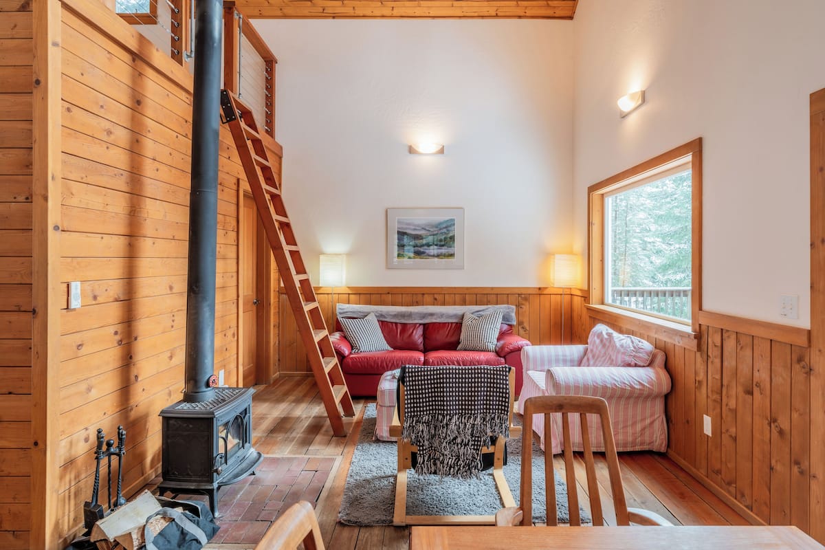 A cozy cabin interior with a wooden interior, a fireplace, a red sofa, a small table, and a window.