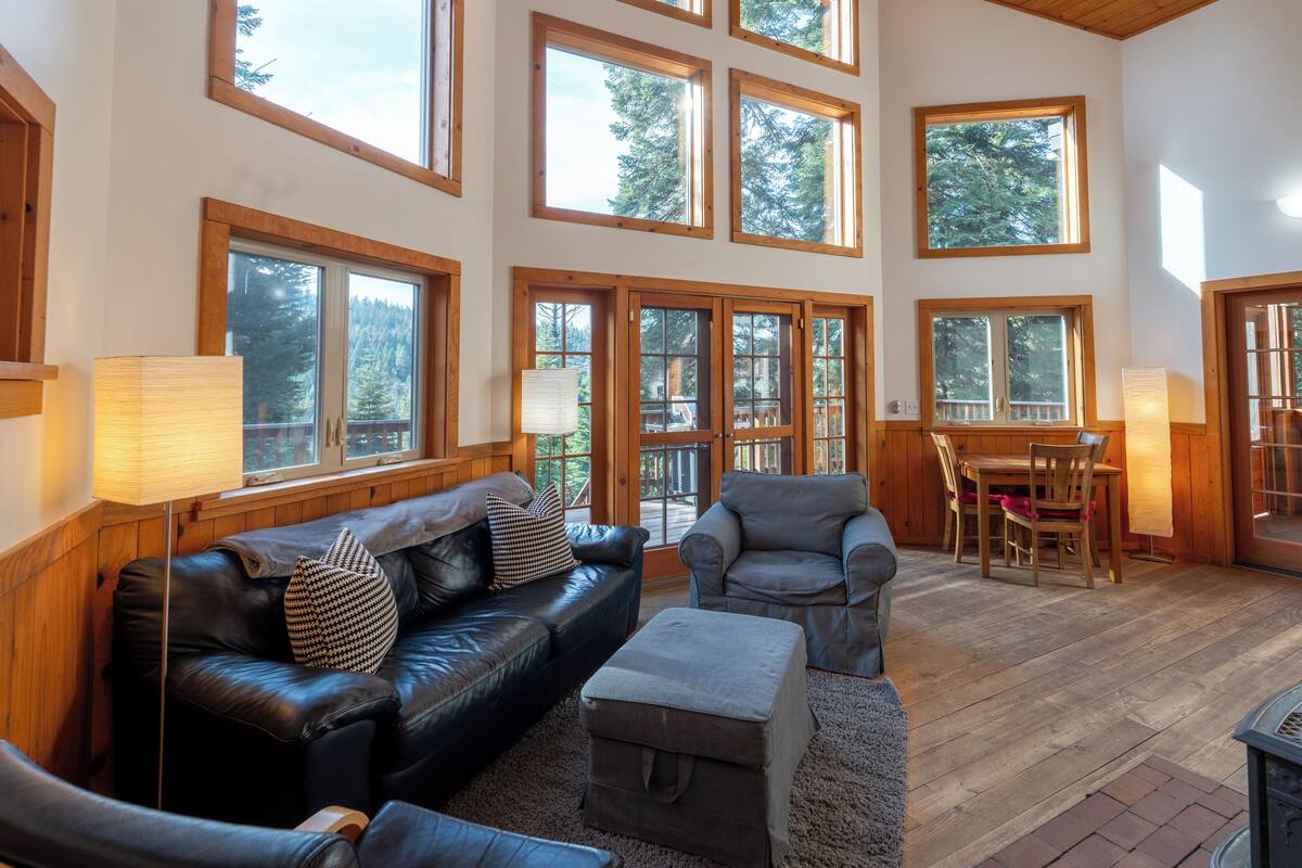 A spacious living room with a leather sofa, a grey armchair, and a wooden dining table. Large windows provide natural light and views of the surrounding forest.
