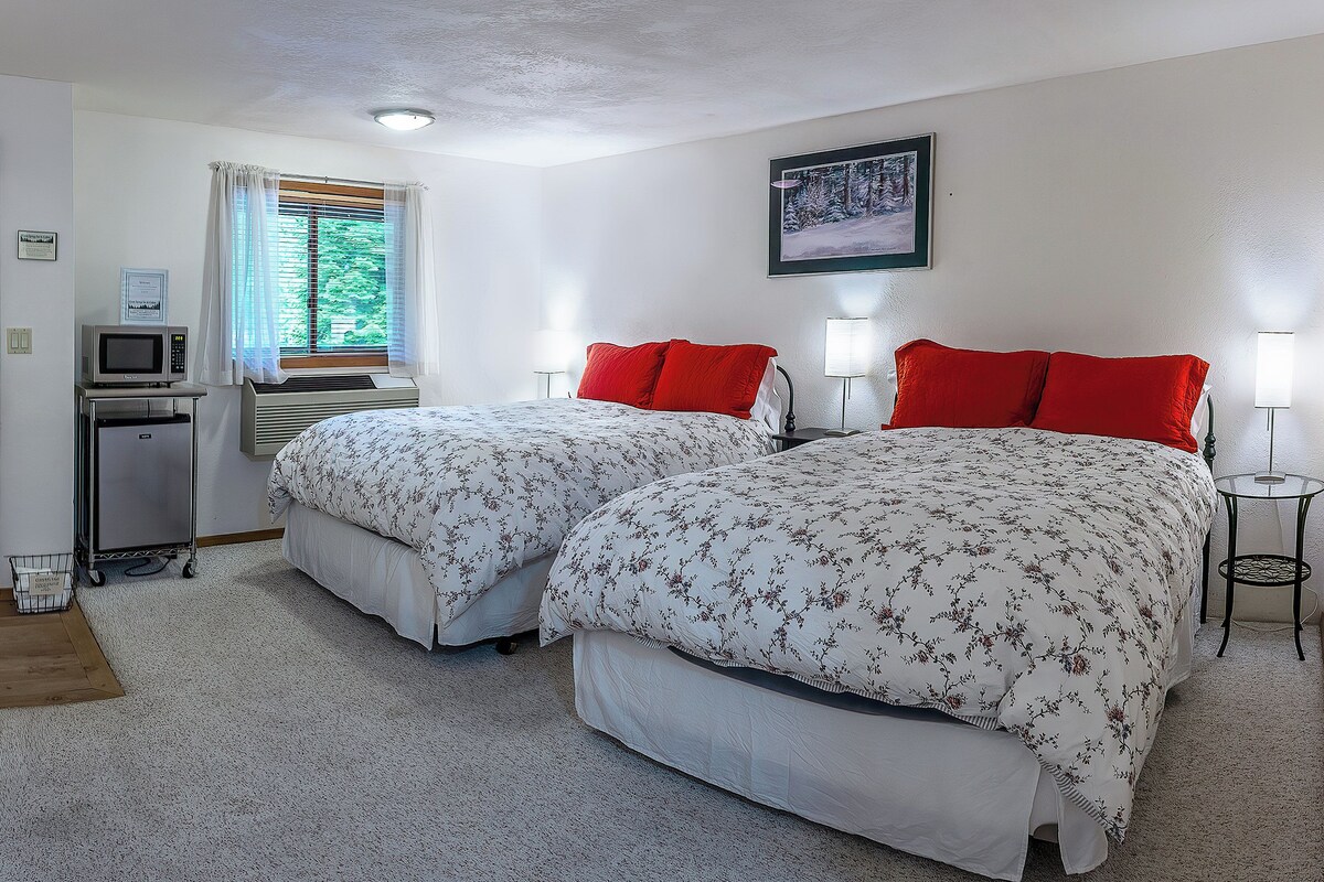 A bedroom with two beds, a desk, a chair, a microwave, and a small table with a lamp.