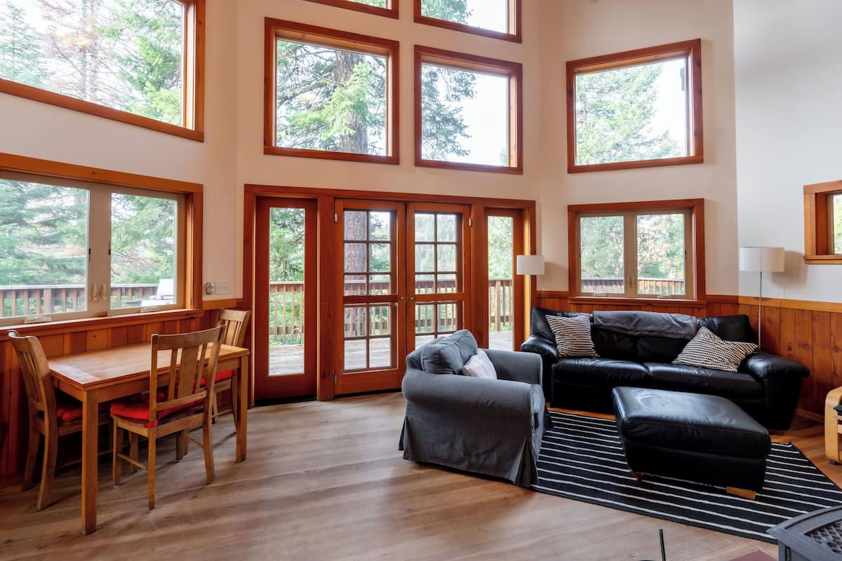 A spacious room with a wooden dining table, chairs, a black leather sofa, and a large window with a view of trees.