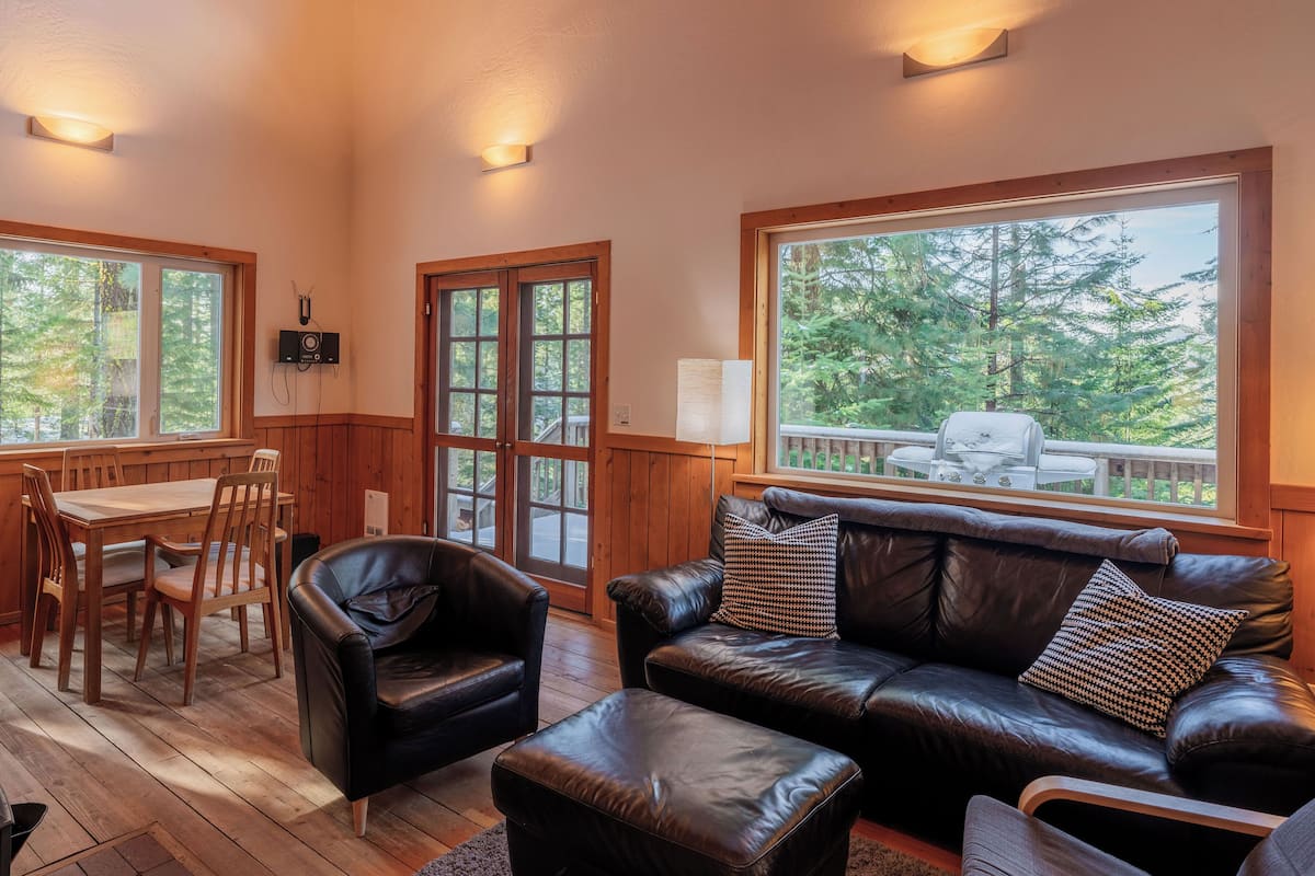 A cozy cabin interior with wooden walls, a leather sofa, a dining table with chairs, and large windows offering a view of the forest.
