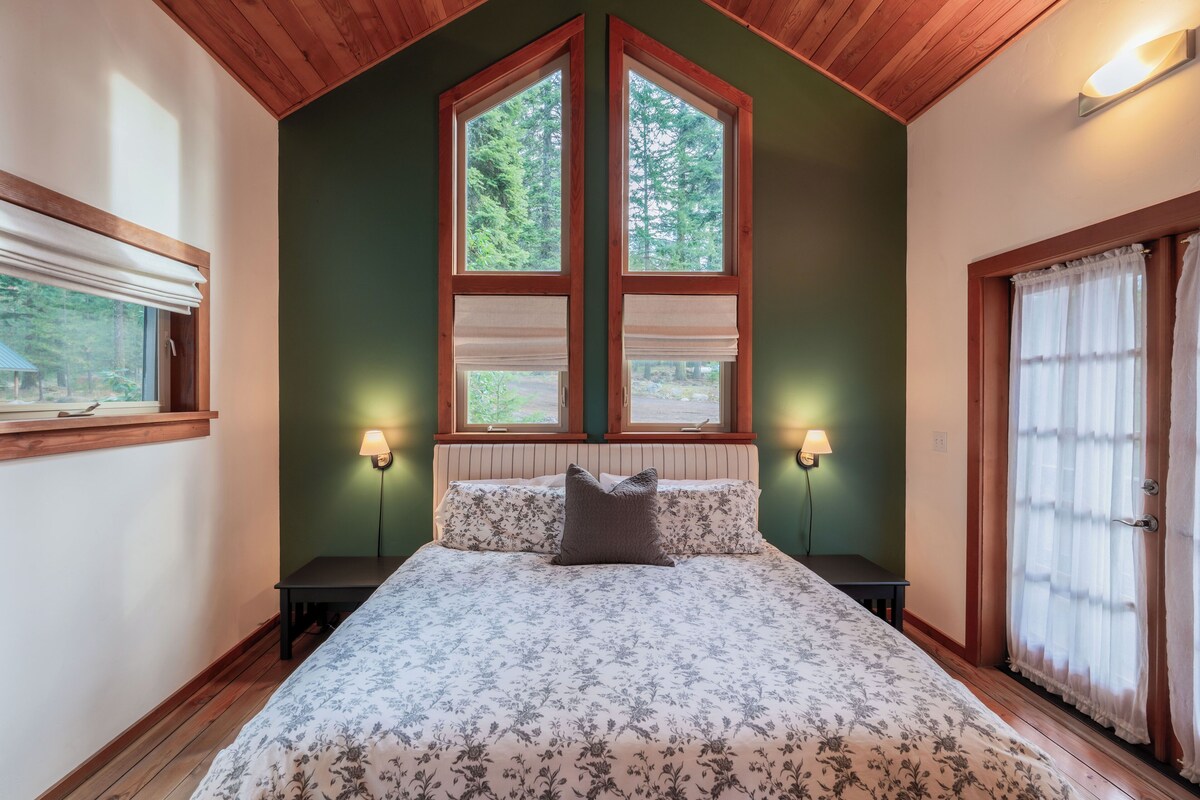 A bedroom with a large bed, two bedside lamps, a window with blinds, and a door with curtains.