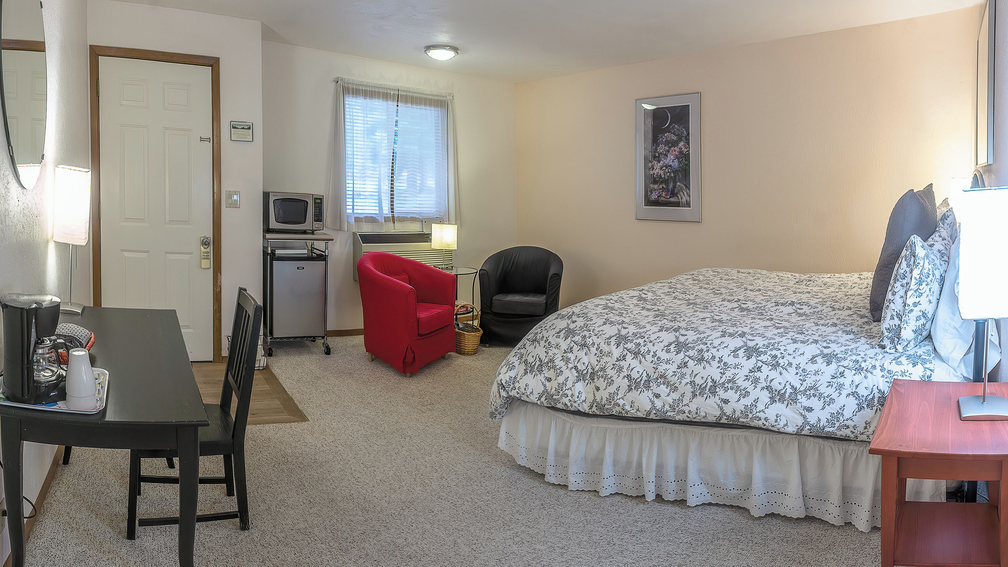 Ein Hotelzimmer mit einem Bett, einem Schreibtisch mit Computer, einem roten Sessel, einem schwarzen Stuhl, einer Mikrowelle und einem gerahmten Bild an der Wand.