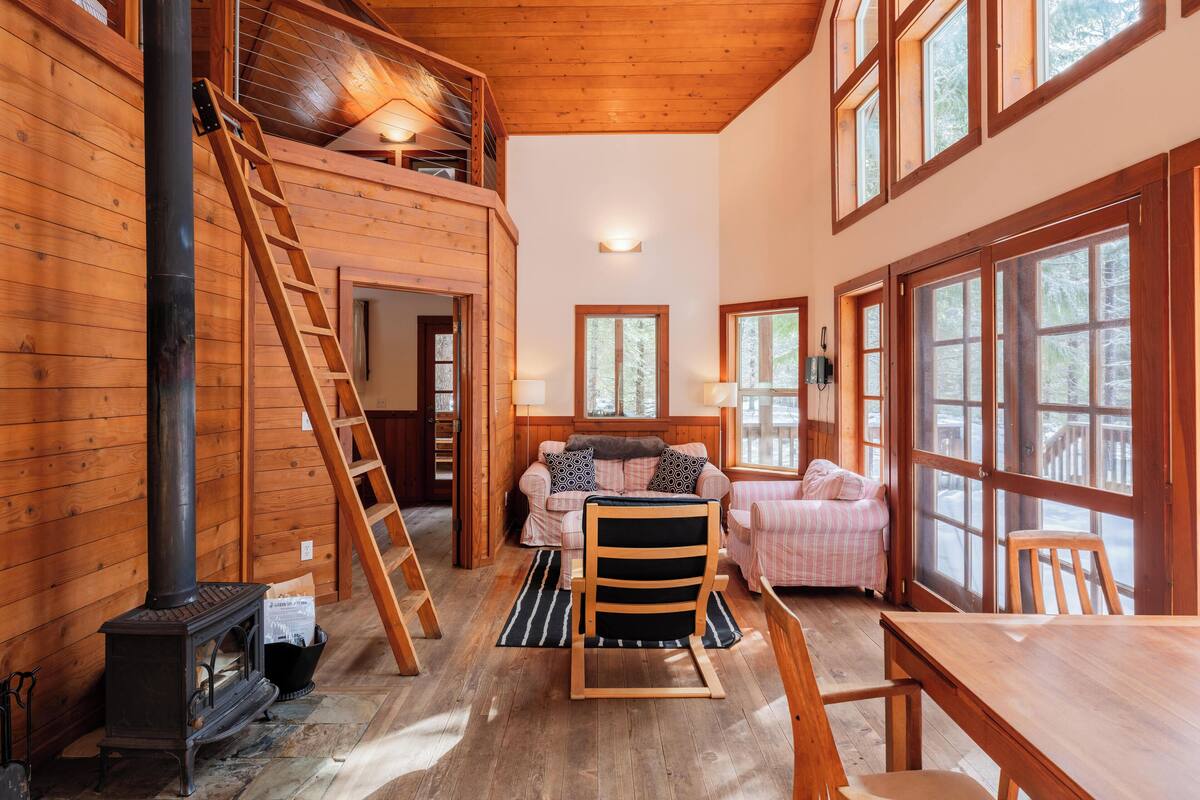 A cozy cabin interior with a loft bedroom, a wooden stove, a dining area, and large windows.