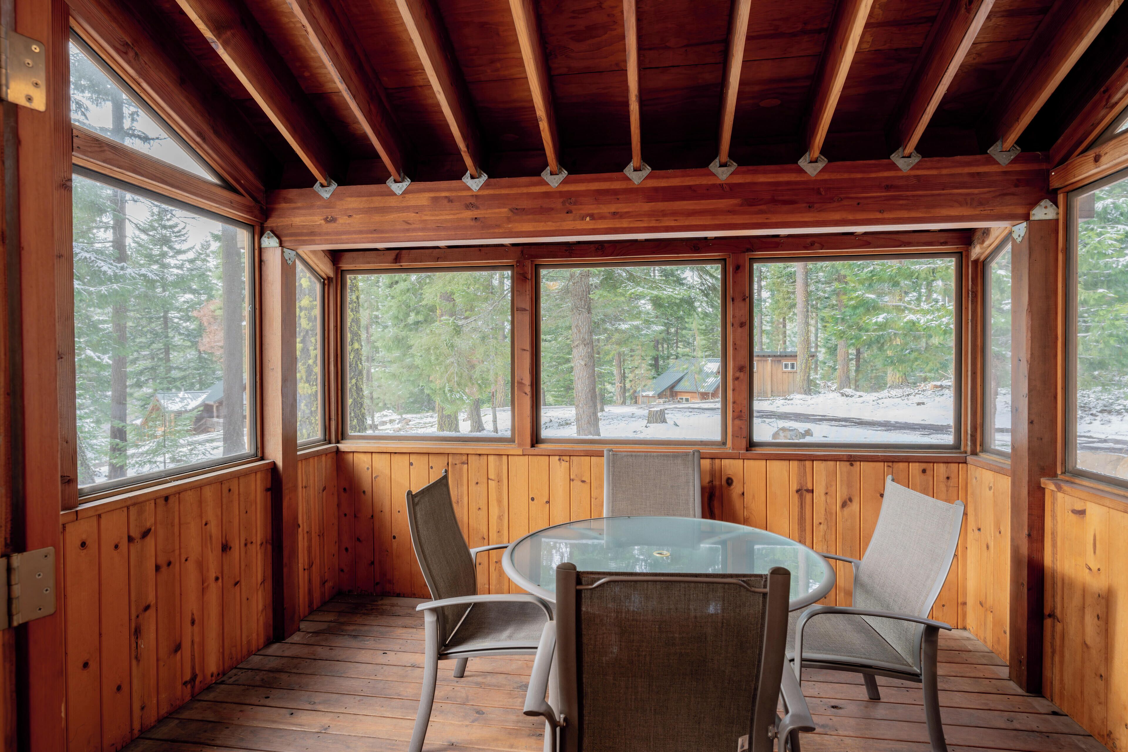 Une cabane en bois avec une table et des chaises en verre, de grandes fenêtres et une vue sur une forêt enneigée.