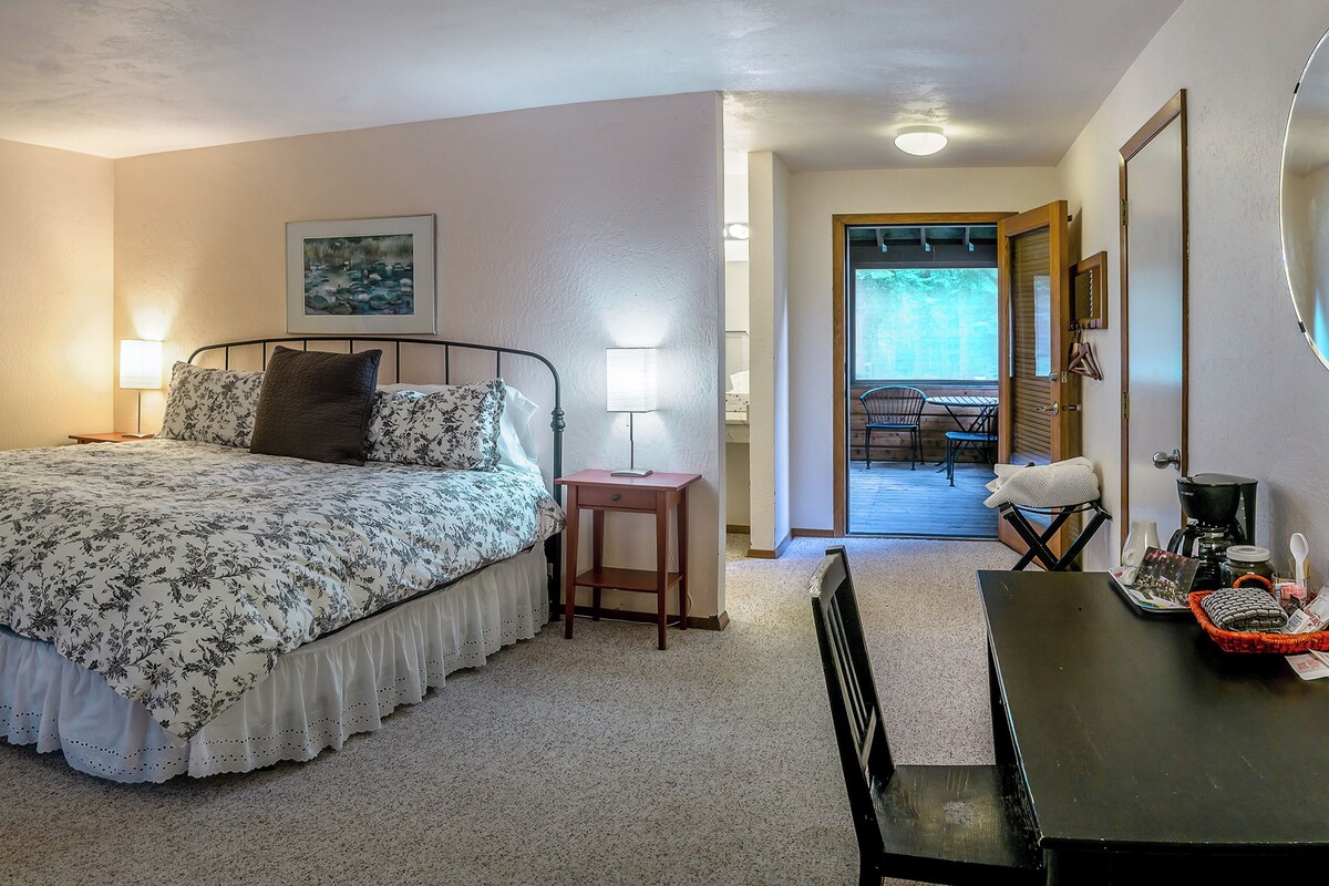 A bedroom with a bed, bedside lamps, a small table, and a chair. There is a door leading to a balcony with outdoor seating.