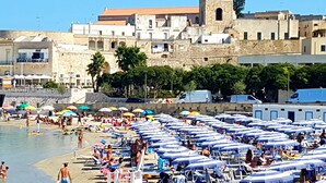 Ein überfüllter Strand mit vielen Sonnenschirmen und Liegestühlen, ein Steinhaus mit einem Turm im Hintergrund und ein klarer, blauer Himmel.
