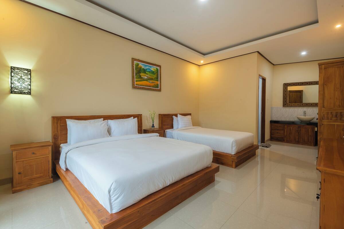 Habitación de hotel con dos camas, una mesita de noche de madera, un tocador, un cuadro, un baño con lavamanos y espejo, y un armario de madera.