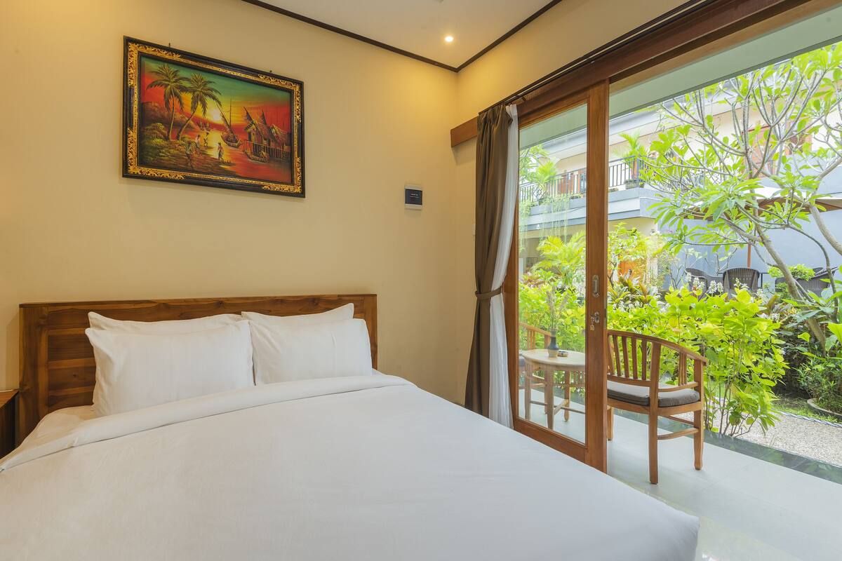 Habitación de hotel con una cama grande, un cuadro en la pared y vistas a un balcón con plantas y una silla.