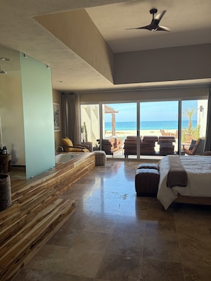 A spacious bedroom with a large bed, a view of the beach, a ceiling fan, and a jacuzzi.