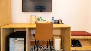 A compact room with a wooden desk, a brown chair, a TV mounted on the wall, and a small bench with shoes. - New Champa Boutique Hotel (Vientiane)