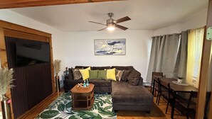A living room with a brown sofa, a wooden coffee table, a patterned rug, a ceiling fan, and a painting on the wall. - Colonial Style Home 5 Br 2 bath n’ Parking! Close to restaurants n’ colleges! (Albany)
