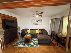 A living room with a brown sofa, a wooden coffee table, a patterned rug, a ceiling fan, and a painting on the wall.