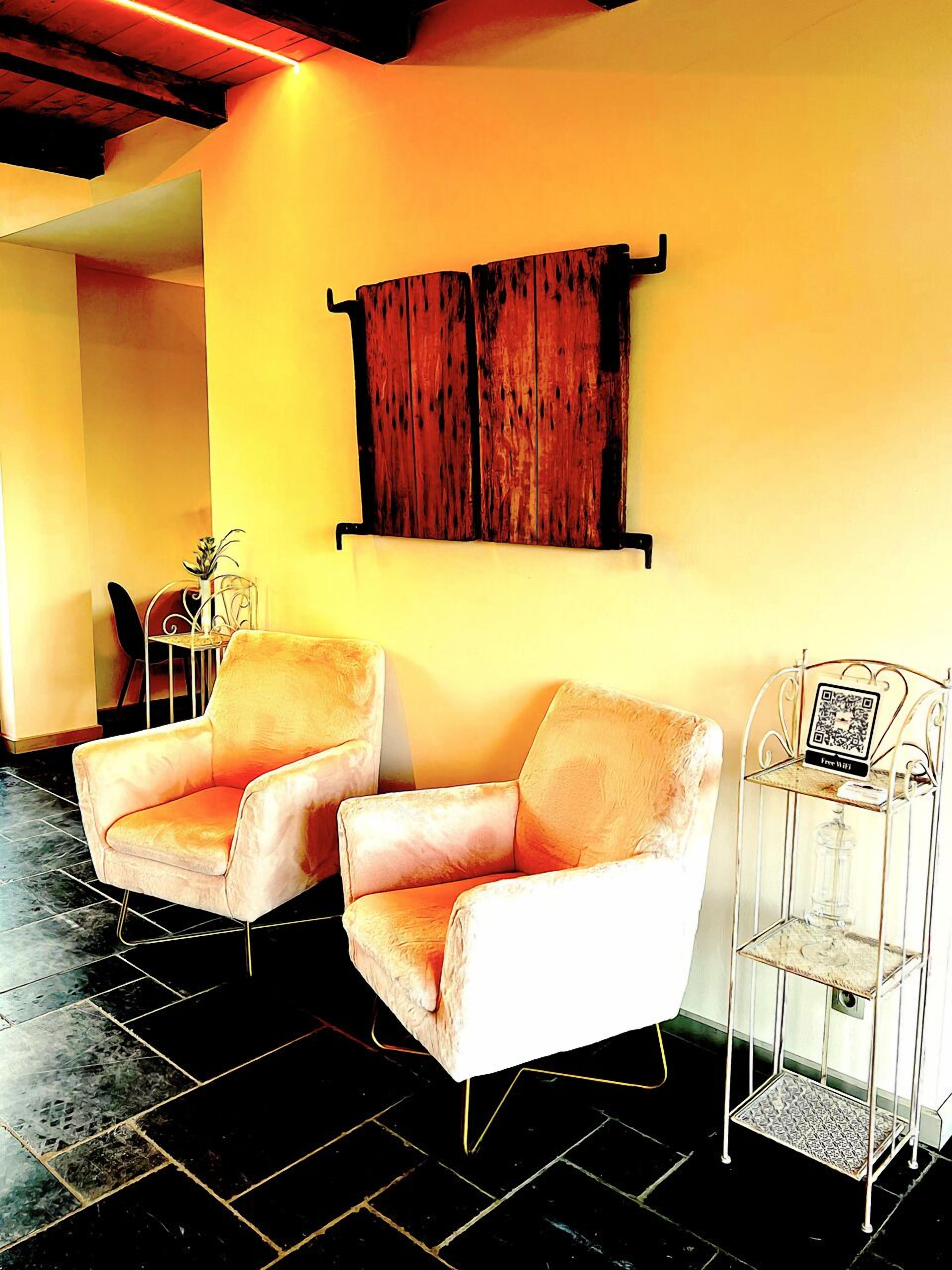 A room with two plush armchairs, a wooden wall art piece, a small table with a vase, and a metal shelf with a QR code.