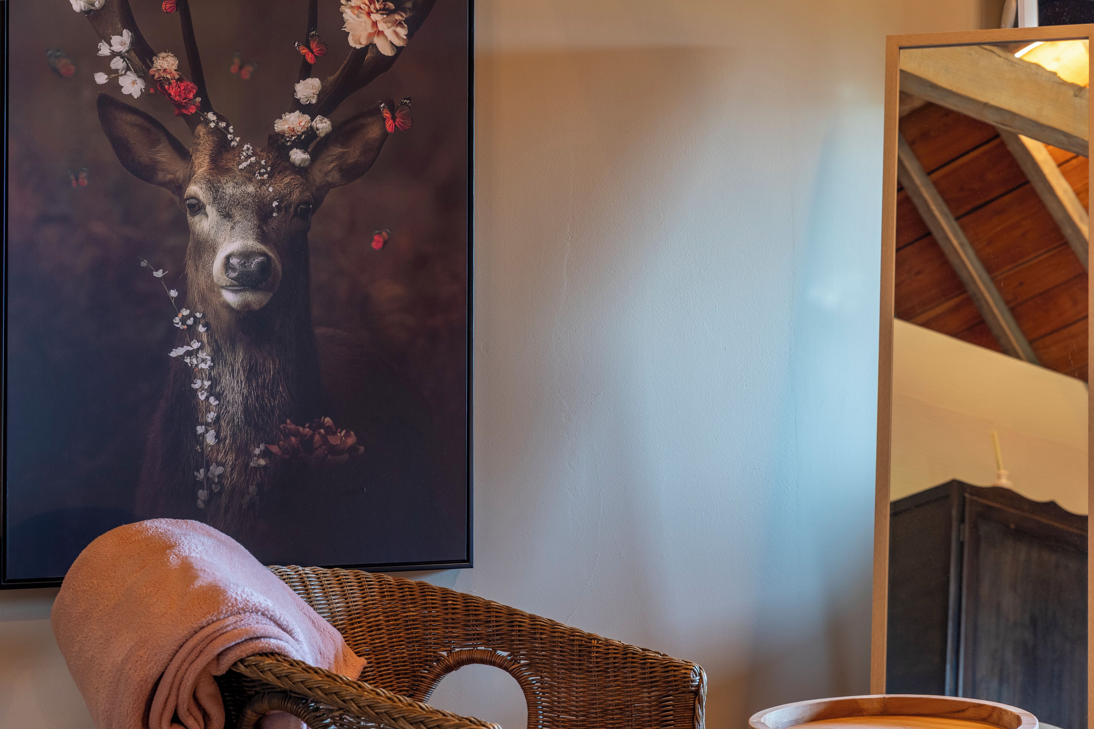 A framed deer painting with flowers and butterflies, a wicker chair with a pink towel, and a wooden ceiling.