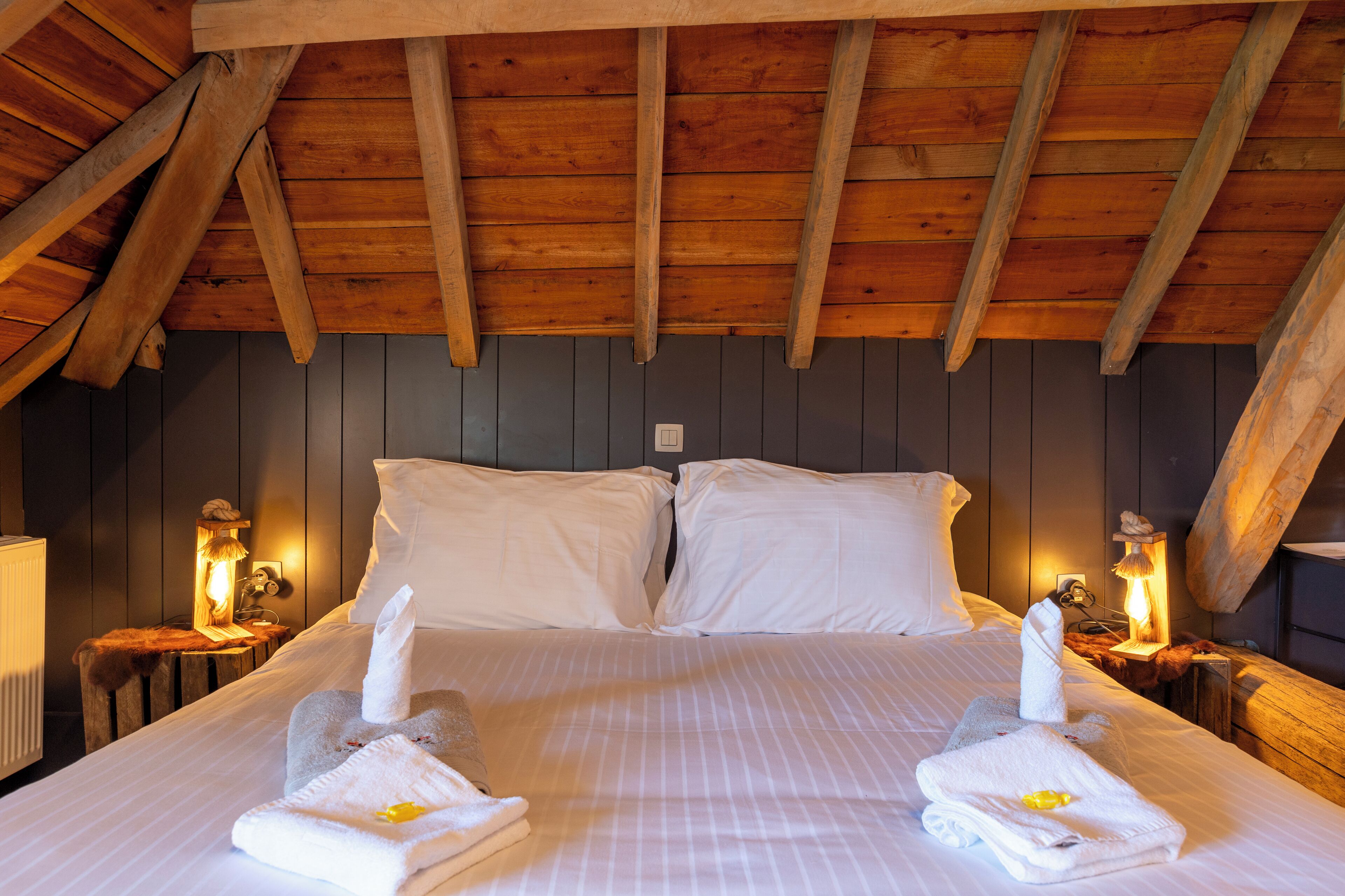 A bedroom with a wooden ceiling, two white beds with pillows, bedside lamps, and towels on the bed.