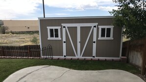Ein kleines, eingeschossiges Gartenhaus mit weißer Tür und Fenstern, umgeben von einer Betonauffahrt und einem Holzzaun.