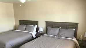 Ein Hotelzimmer mit zwei Betten, jeweils mit grauer Bettwäsche und Holz-Kopfteilen. Auf der einen Seite befindet sich ein Nachttisch mit einer Lampe und einer Taschentuchbox.