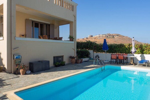 A swimming pool with a view of a hill, outdoor furniture, and potted plants.