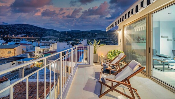 A balcony with sun loungers, a glass door leading to an interior room, and a view of the cityscape and mountains in the background.