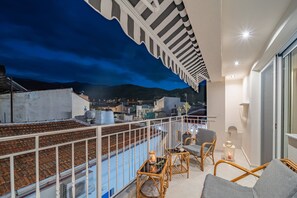 Varanda com vista para a cidade ao entardecer, mobiliário de vime e guarda-corpo branco.