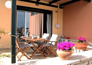 An outdoor patio with a wooden table and chairs, a potted plant with pink flowers, and a wall-mounted air conditioning unit. - Nina's Place, 5min On Foot From The Beach (Panormos)