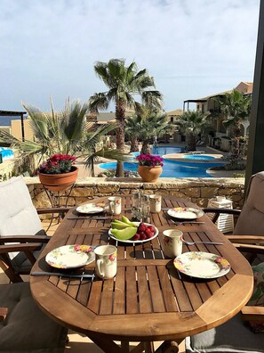Une table en bois, dressée pour le repas, avec des assiettes, des tasses et un saladier de fruits, donnant sur une piscine bordée de palmiers et sur un bâtiment en arrière-plan.
