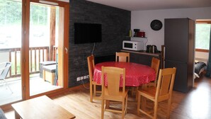 A cozy living room with a round table covered in a red polka dot tablecloth, wooden chairs, a flat-screen TV mounted on a dark brick wall, a microwave, and a refrigerator.