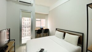 A hotel room with a bed, a desk with two chairs, an air conditioner, and a window with a view of buildings. - Chic And Cozy Studio Apartment At Transpark Juanda Bekasi (Bekasi)