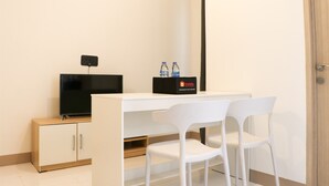 A small, modern room with a white desk, two white chairs, a flat-screen TV, a black box with water bottles, and a wooden cabinet.