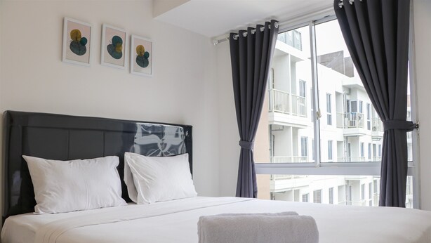A neatly made bed with white linens and pillows, a grey towel on the bed, and a window with grey curtains.