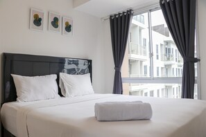 A neatly made bed with white linens and pillows, a grey towel on the bed, and a window with grey curtains. - Cozy And Enjoy Living 2Br At Tokyo Riverside Pik 2 Apartment (Teluknaga)